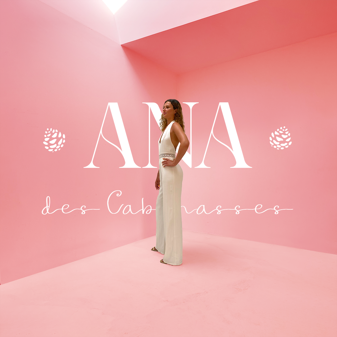 Dans l'élégance intemporelle d'un espace minimaliste peint en rose, notre modèle porte une combinaison de mariage sophistiquée, incarnant l'essence d'Ana des Cabanasses. La conception du logo présente 'Ana' dans le style art déco français, 'des Cabanasses' dans une écriture gracieuse, et deux pommes de pin, évoquant subtilement l'origine de la marque parmi les pins du sud-ouest.